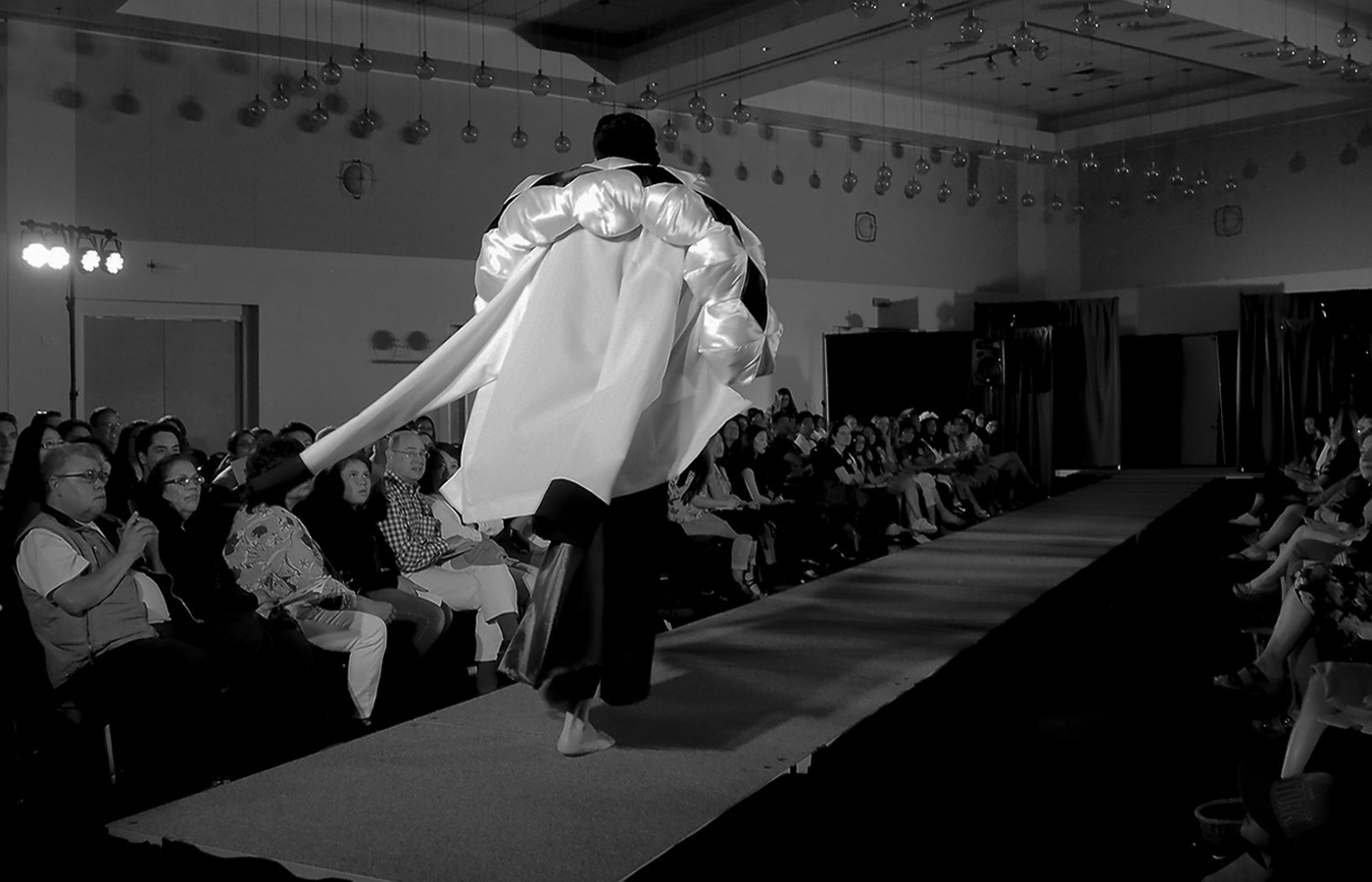 A black and white image of a figure walking along a long fashion runway with an audience of people on either side of it. The model wears a white coat with a large stuffed boa-shaped neckpiece with a diamond motif.